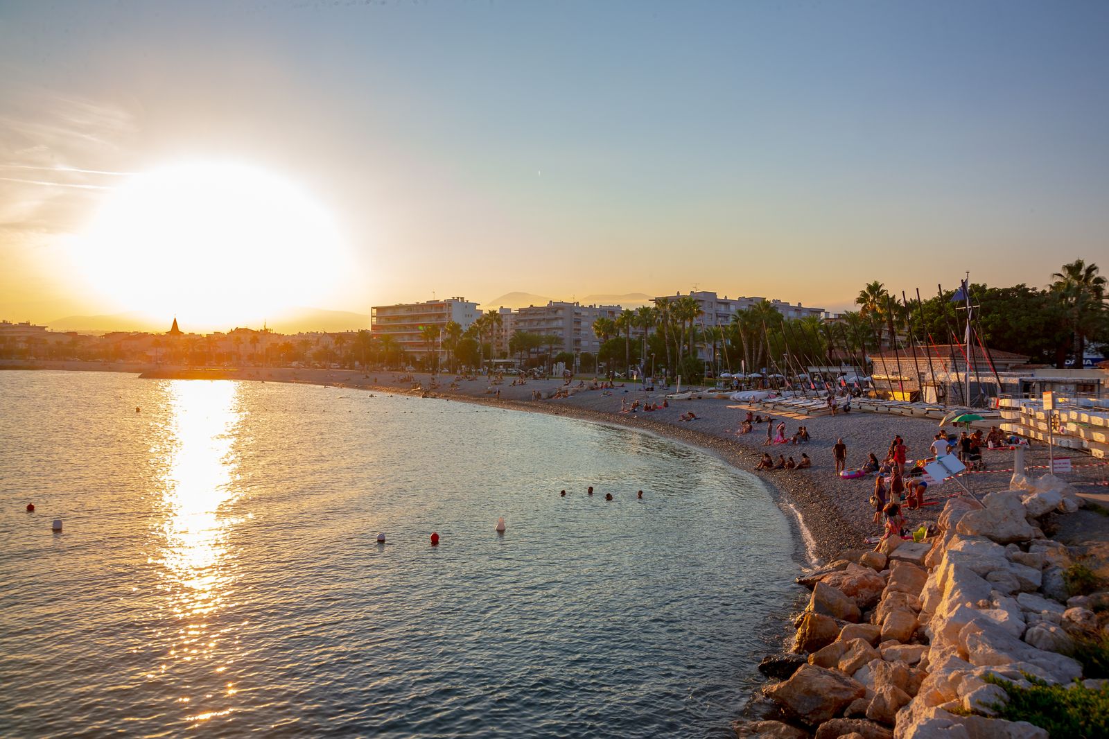 The pebble beach and seafront of Cros-de-Cagnes at sunset, with swimmers in the bay and sailing dinghies drawn up along the shore
