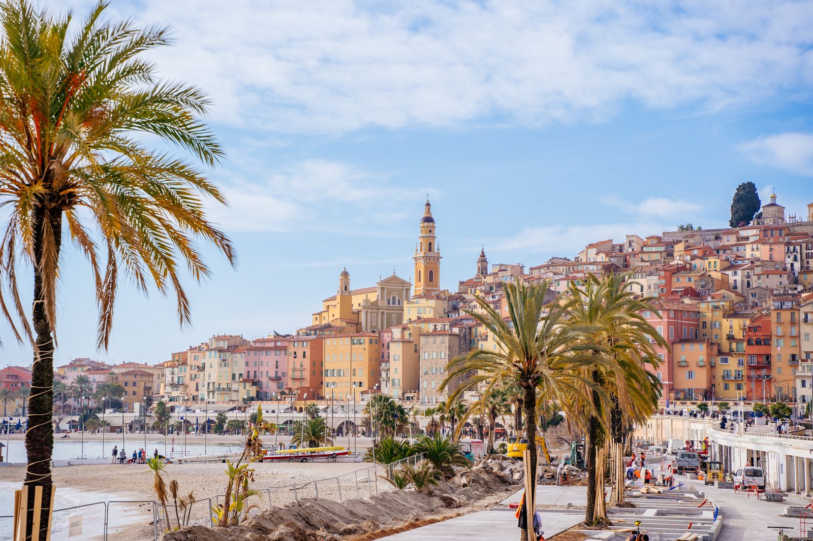 The old town of Menton rising above the beach in ochre, yellow and rose, with the Basilica of Saint-Michel and palm trees along the seafront