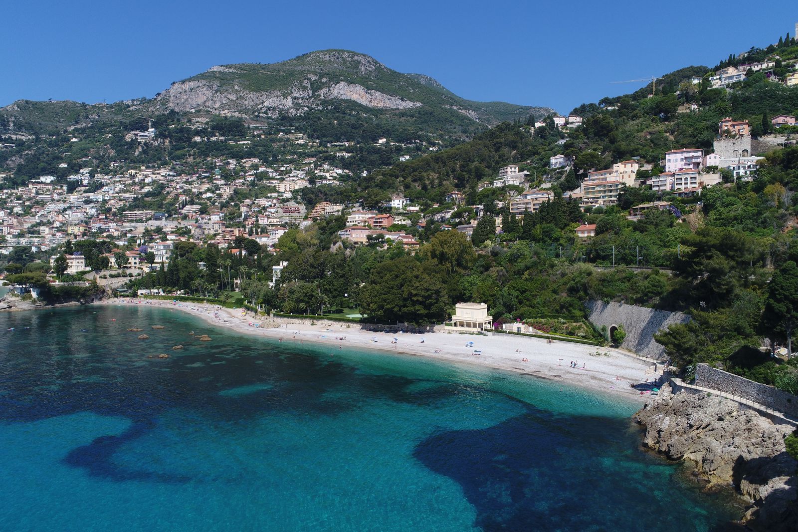 Aerial view of Plage de la Buse and the wooded slopes of Cap Martin, with hillside villas rising behind the pebble beach and turquoise water