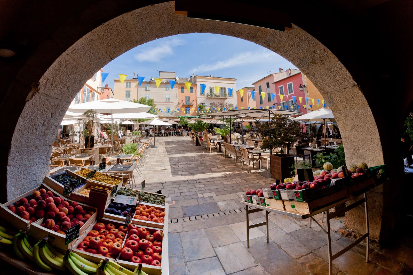 Place des Arcades in Valbonne seen through a stone arch, with market fruit stalls in the arcade, bunting overhead and cafe terraces filling the square
